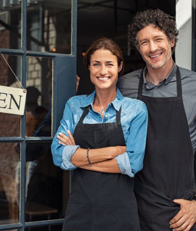 Two cheerful small business owners smiling and looking at camera while standing at entrance door. Happy mature man and mid woman at entrance of newly opened restaurant with open sign board. Smiling couple welcoming customers to small business shop.