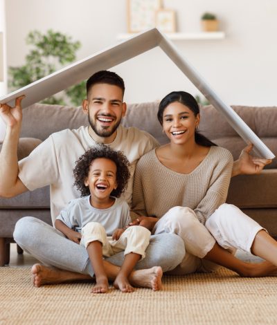 Cheerful parents with child smiling and keeping roof mockup over heads while sitting on floor in cozy living room during relocation